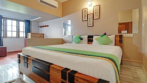 Une chambre d’hôtel avec un grand lit, une tête de lit en bois, une fenêtre avec des rideaux, un climatiseur fixé au mur et des tableaux encadrés accrochés au mur.