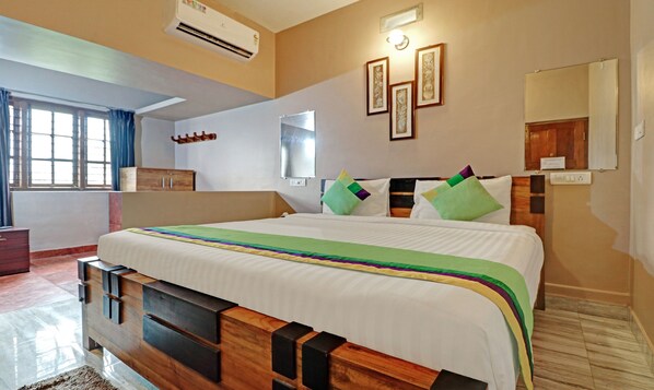 A hotel room with a large bed, a wooden headboard, a window with curtains, a wall-mounted air conditioner, and framed pictures on the wall.