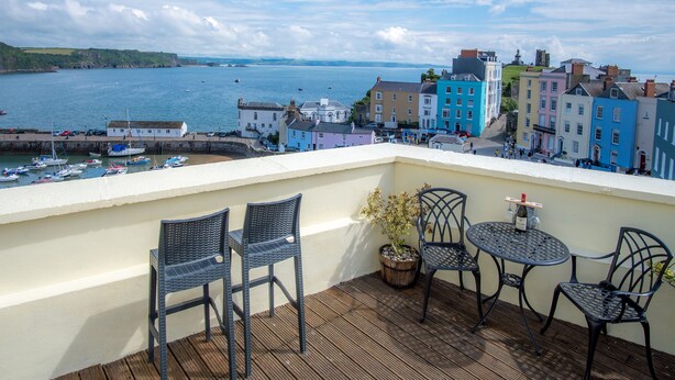 Un balcon agrémenté de fauteuils et d’une table en osier, donnant sur un port où se trouvent des bâtiments aux couleurs vives et des bateaux.