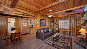 A rustic cabin interior with a wooden floor, a leather sofa, a dining table, a kitchen area, and a staircase.