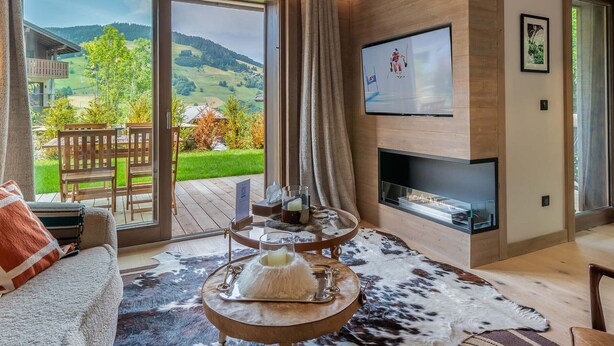 A modern living room with a fireplace, a television, and a scenic view of a mountainous landscape through large sliding doors. - Charming Garden-Level Apartment in Rochebrune, Megève – Sleeps 6 (Megève)