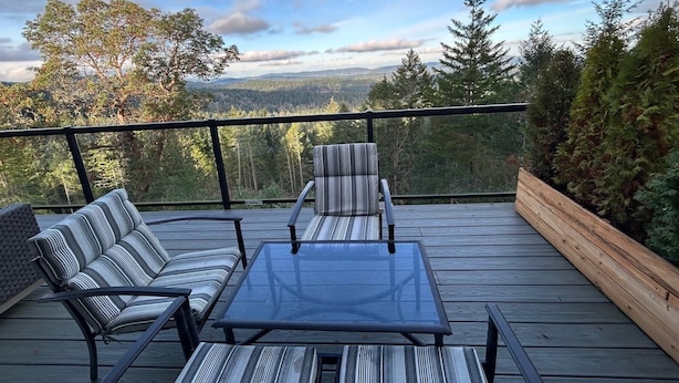 A deck with a glass-top table and striped chairs, overlooking a forested landscape. - Entire Suite, Ocean, mountain view, hot tub, next to Mountain Maxwell park. (Salt Spring Island)