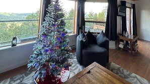 A cozy living room with a wooden ceiling, a large window with a view of trees, a Christmas tree, a dark sofa, and a small wooden table. - Entire Suite, Ocean, mountain view, hot tub, next to Mountain Maxwell park. (Salt Spring Island)
