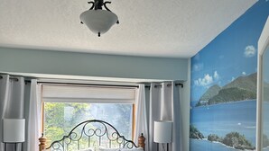 A bedroom with a bed, a window with curtains, a ceiling fan, and a mural of a coastal scene. - Entire Suite, Ocean, mountain view, hot tub, next to Mountain Maxwell park. (Salt Spring Island)