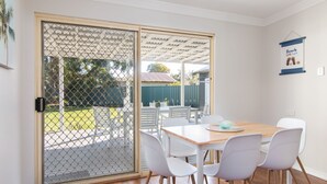 A dining area with a wooden table and white chairs, a sliding glass door leading to a backyard, and a wall-mounted sign with beach-related text. - 9 Breckenridge Street (Forster)