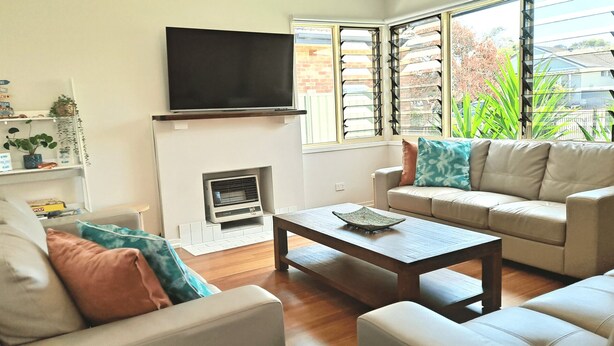 A living room with a flat-screen TV mounted above a fireplace, a beige sofa with cushions, a wooden coffee table, and a window with blinds. - 9 Breckenridge Street (Forster)
