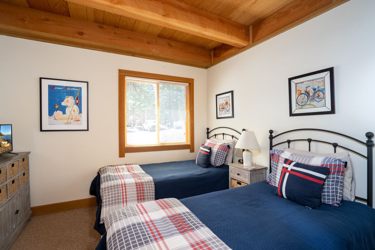 A bedroom with two beds, a wooden ceiling, a window with blinds, and framed pictures on the walls.