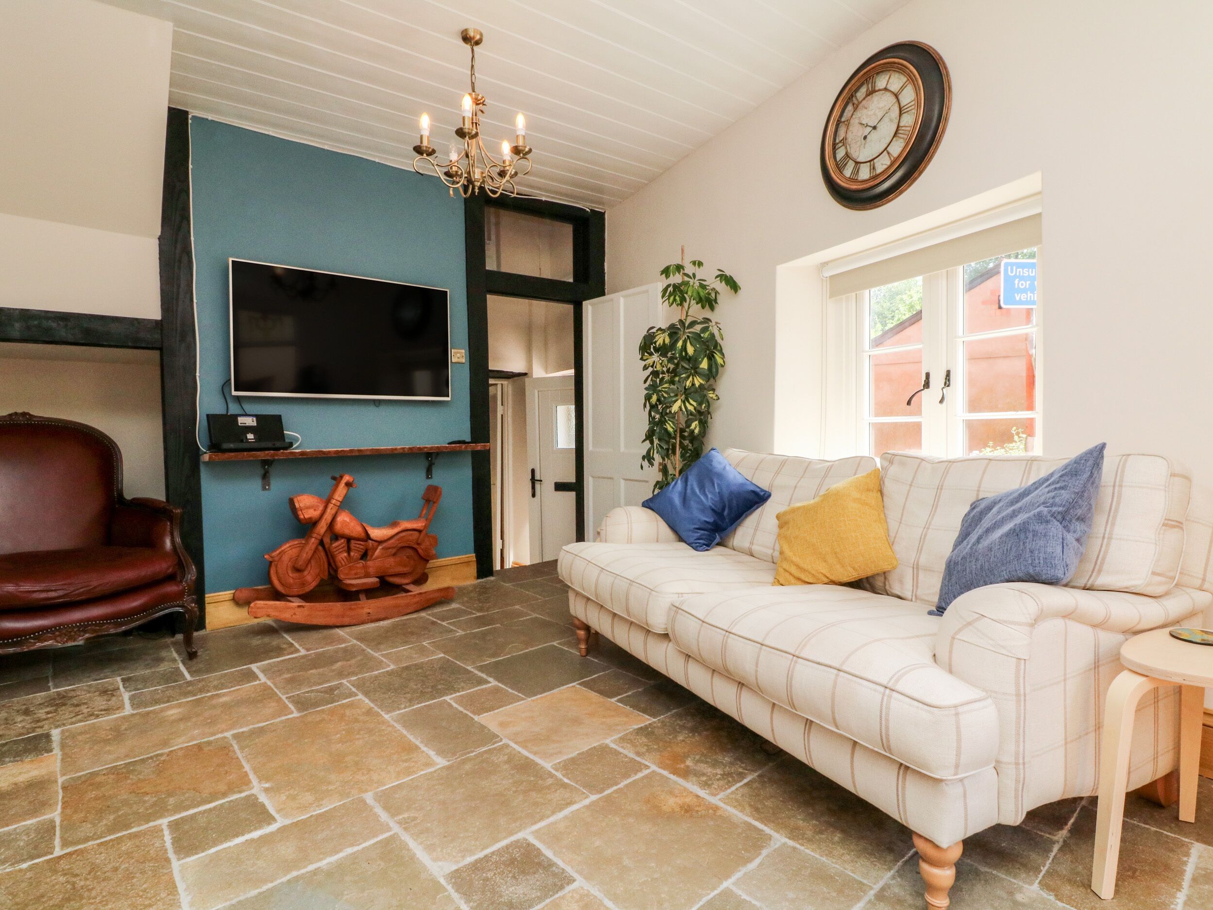 A living room with a large flat-screen TV, a brown leather chair, a white sofa with colorful cushions, a wooden rocking horse, a chandelier, and a clock on the wall.