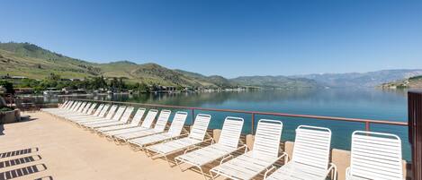 A deck with white lounge chairs facing a calm lake, surrounded by hills.