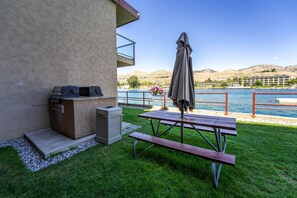 A patio with a grill, picnic table, and umbrella overlooking a lake with mountains in the background.