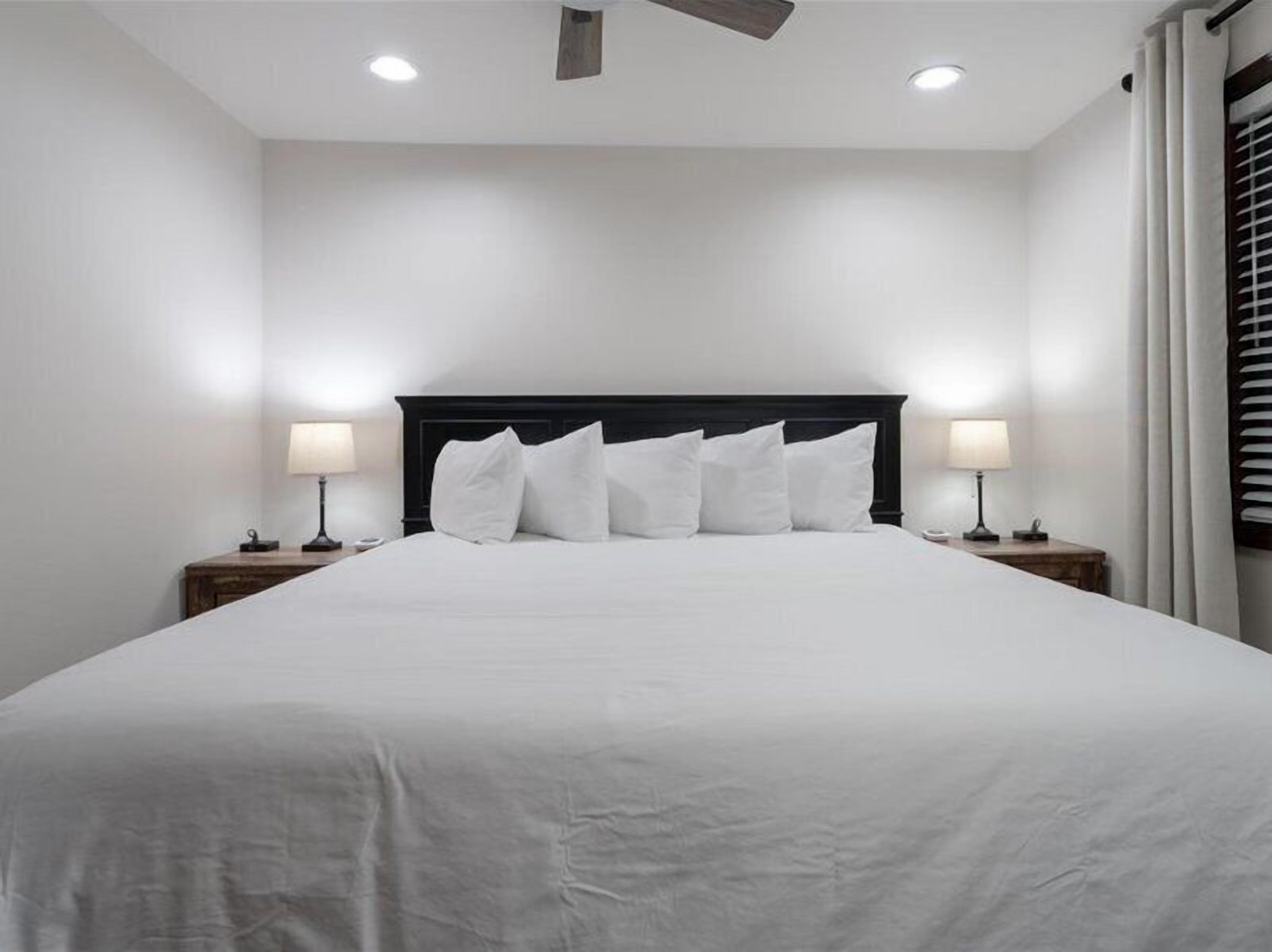 Una cama bien hecha con sábanas blancas y un cabecero oscuro en una habitación con paredes blancas y dos mesitas de noche con lámparas.