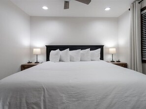 A neatly made bed with white linens and a dark headboard in a room with white walls and two bedside tables with lamps.