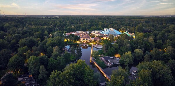 An aerial view of a hotel surrounded by dense forest, featuring a large glass-roofed building, a dock, and a small boat. - Hotel De Eemhof by Center Parcs (Zeewolde)