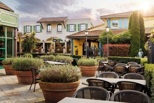 Un café al aire libre con mesas y sillas, rodeado de plantas en macetas y edificios con arquitectura tradicional.