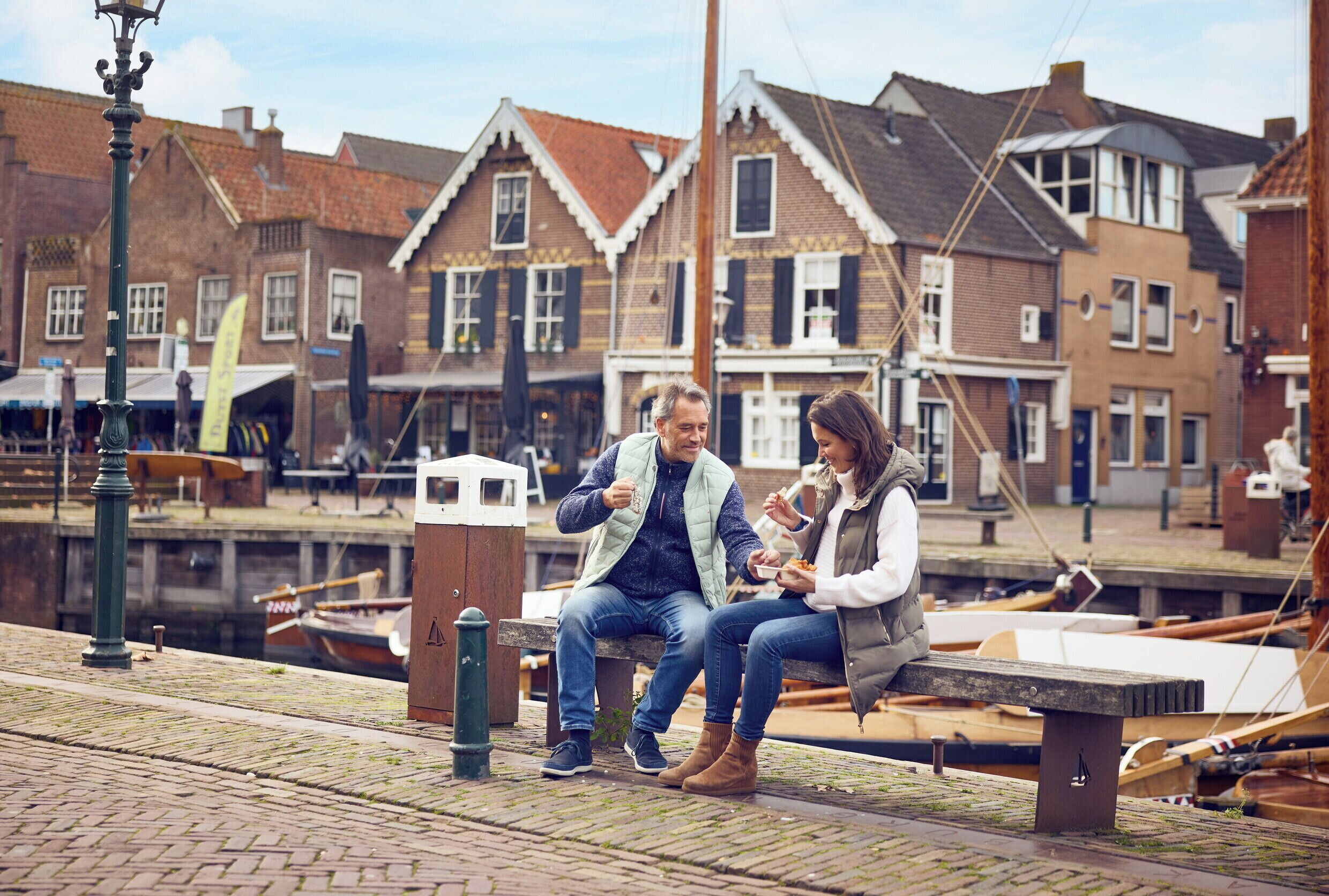 Una pareja sentada en un banco junto a un canal, con edificios tradicionales al fondo.