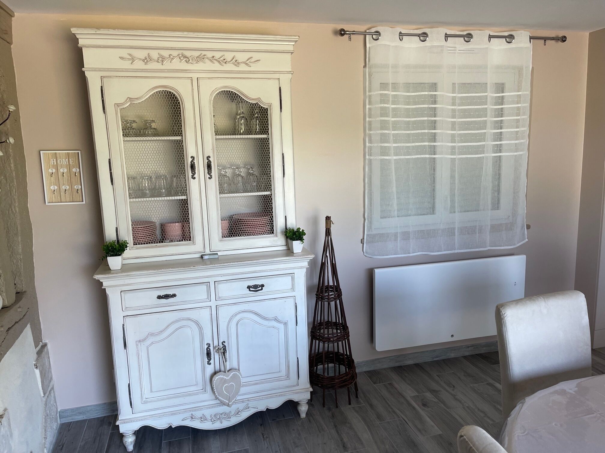 Une pièce avec un meuble blanc aux portes vitrées, une chaise en osier et un radiateur.