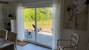 Ein Zimmer mit einer Schiebetür zu einem Außenbereich mit Sitzgelegenheiten und einem Kiesweg. Im Zimmer steht ein weißer Sessel, ein kleiner Tisch und ein Bild an der Wand.