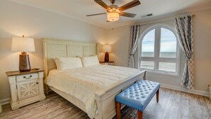 A bedroom with a bed, bedside lamps, a bench, a ceiling fan, and a view of the ocean through a window. - Oceanfront 2BR condo at Villa Capriani 317A, North Topsail Beach, perfect for families. (North Topsail Beach)