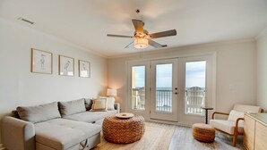 A living room with a grey sofa, a round woven ottoman, a small table with a lamp, and a view of the ocean through a sliding glass door. - Oceanfront 2BR condo at Villa Capriani 317A, North Topsail Beach, perfect for families. (North Topsail Beach)