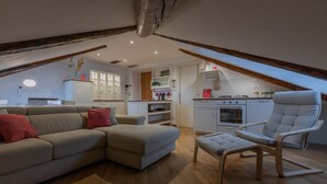 Un salon chaleureux avec un canapé, un pouf et un fauteuil à haut dossier. On aperçoit une cuisine avec des meubles blancs et une cuisinière au fond.