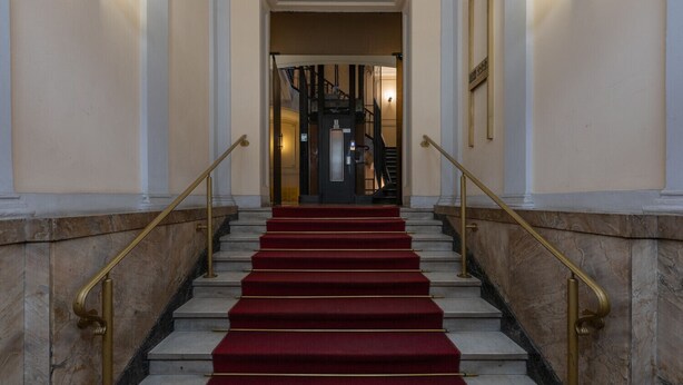 Un magnifique escalier, avec des marches recouvertes de moquette rouge et des rampes dorées, menant à un intérieur sombre.