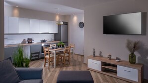 A modern kitchen with white cabinets, a dining area with wooden chairs, a flat-screen TV mounted on the wall, and a small side table with decorative items. - Archipelagos Apt with Unique Sea & Old Port View in the City Center (Heraklion)