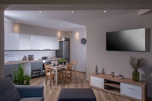 A modern kitchen with white cabinets, a dining area with wooden chairs, a flat-screen TV mounted on the wall, and a small side table with decorative items. - Archipelagos Apt with Unique Sea & Old Port View in the City Center (Heraklion)