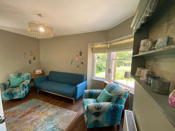 A cozy living room with a blue sofa, patterned armchairs, a decorative light fixture, and a view of greenery through the window.