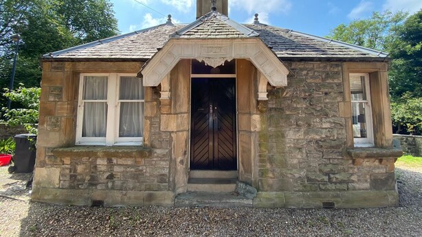 A stone cottage with a gabled roof, a central entrance with a black door, and a small porch area.