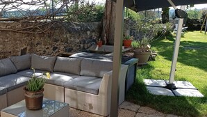 A covered outdoor seating area with a grey sofa and a table, surrounded by potted plants and a stone wall. - Un Lieu Bucolique, Idéal Pour se Ressourcer (Beauzac)