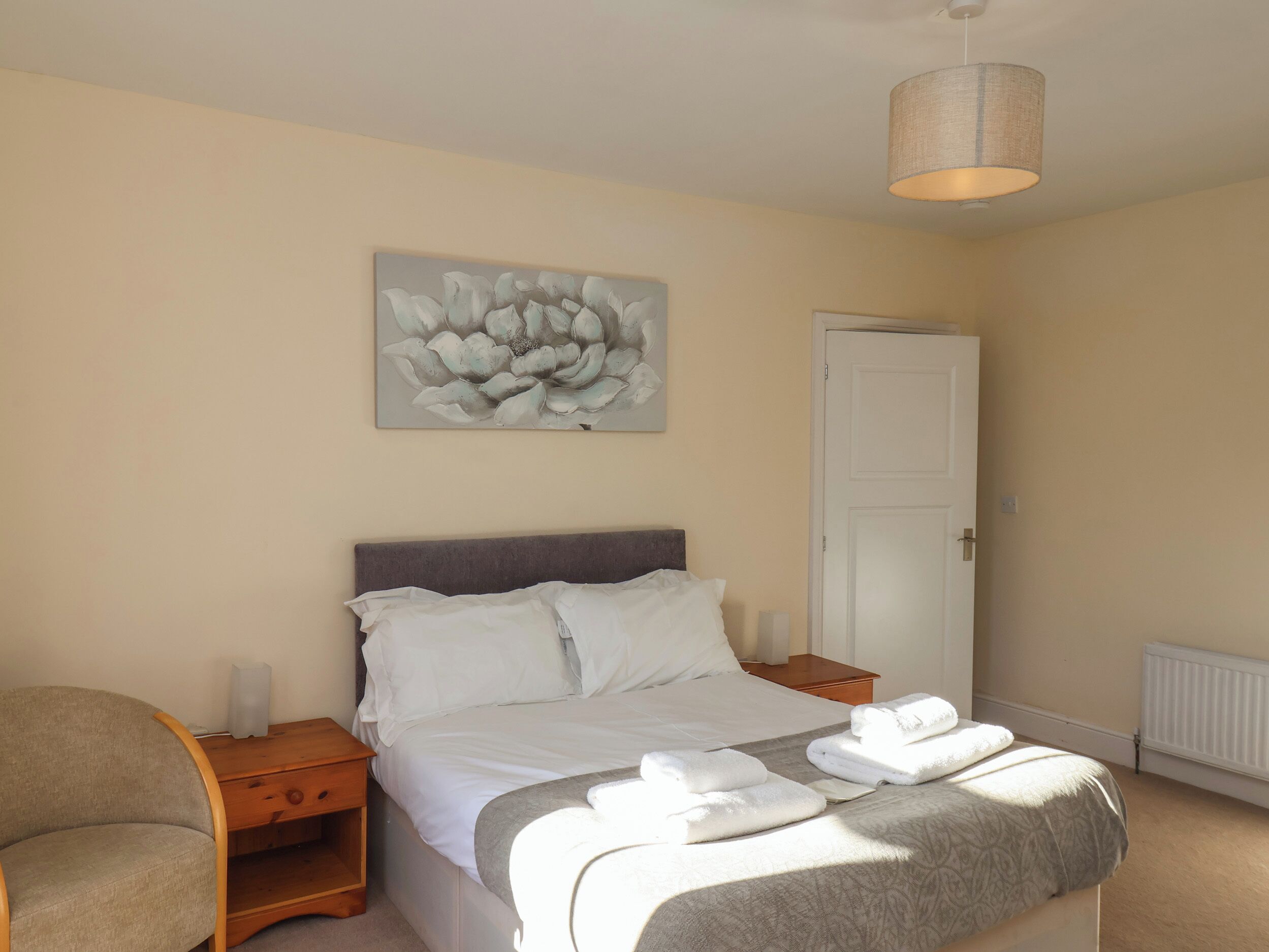 A neatly made bed with white linens and a grey throw blanket, a bedside lamp, and a wall-mounted artwork of a flower.