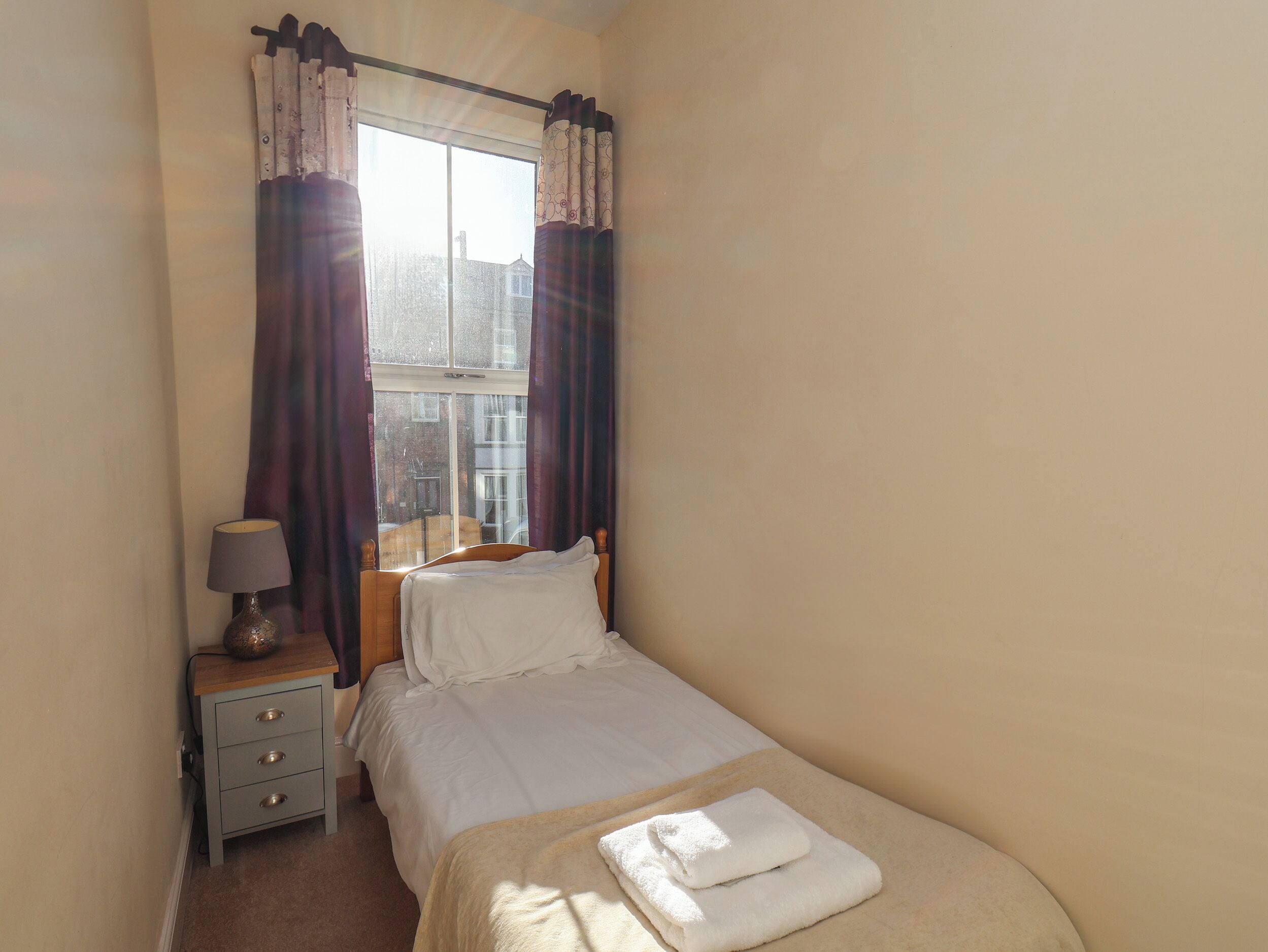 A small, neatly made bedroom with a single bed, a bedside table with a lamp, and a window with curtains.
