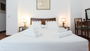 Una cama bien hecha con sábanas y toallas blancas, dos lámparas de noche, un cuadro en la pared y una silla de madera con un cojín verde.