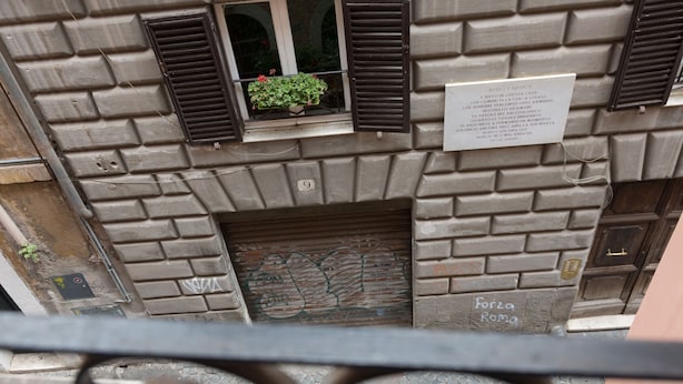 Fachada de un edificio con una ventana que muestra una planta en maceta, una placa con texto y una puerta que dice "Forza Roma".