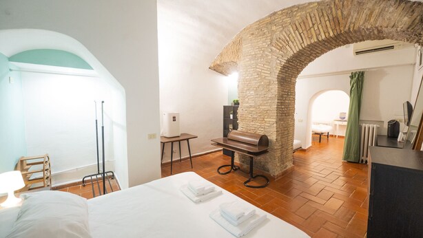 A hotel room with a bed, a desk, a chair, a stone archway, and a view of the dining area. - Campo de Fiori Grotto Apartment (Roma)