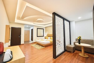 Habitación de hotel con cama, zona de estar con sofá y una mesita, escritorio de madera con teléfono y bandeja con tazas, y un separador decorativo con estampado floral.