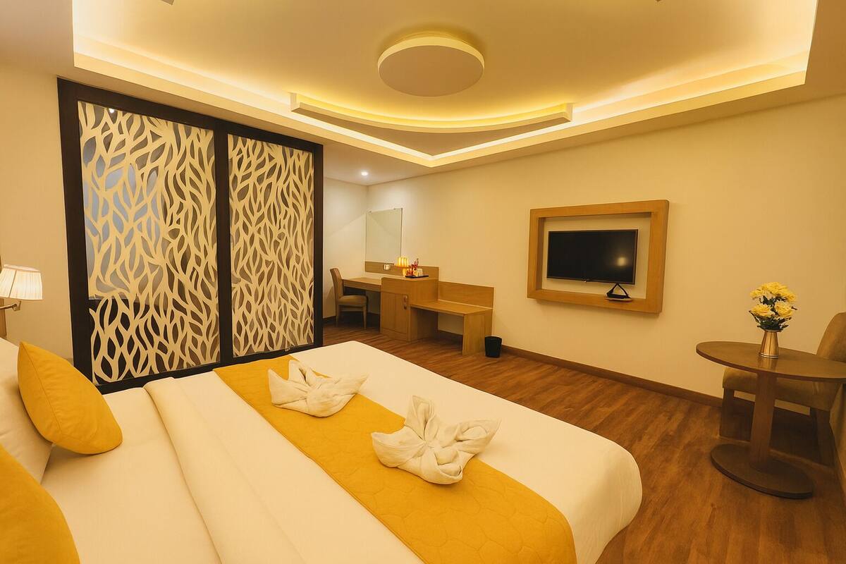 A hotel room with a bed, a desk, a chair, a TV, and a small table with flowers.