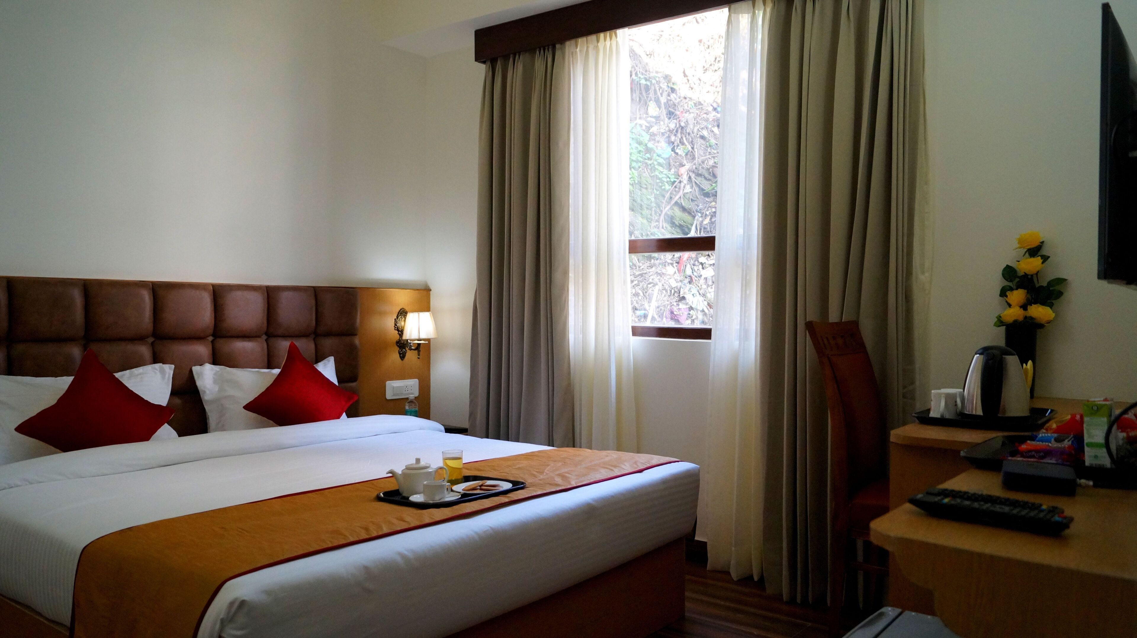 A hotel room with a large bed, a wooden headboard, a bedside table with a lamp, a window with curtains, and a desk with a remote control.