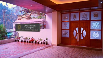 Entrada de hotel con un cartel, plantas en macetas y una puerta de madera con paneles de vidrio.