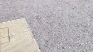 Un camino de grava conduce a un portón de piedra, rodeado de vegetación exuberante y colinas al fondo.