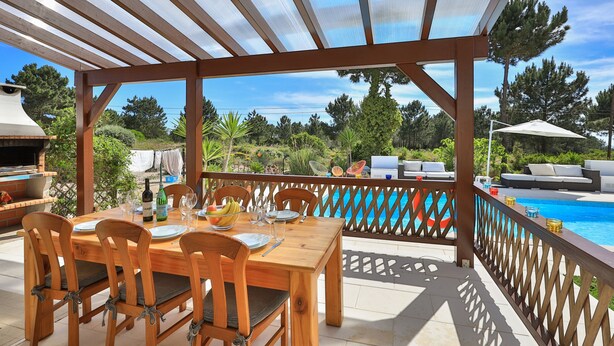 Eine Terrasse im Freien mit Holztisch und -stühlen, einem Grill und Blick auf einen Swimmingpool.