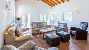 Ein großzügiges Wohnzimmer mit hoher Holzbalkendecke, großen Fenstern und vielfältigen Sitzmöglichkeiten, darunter eine Couch, Sessel und Poufs.