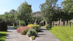 A stone pathway leading to a historic building with a tower, surrounded by lush greenery and flowering plants.