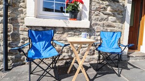 Two blue folding chairs and a small wooden table outside a stone building with a window displaying red flowers.