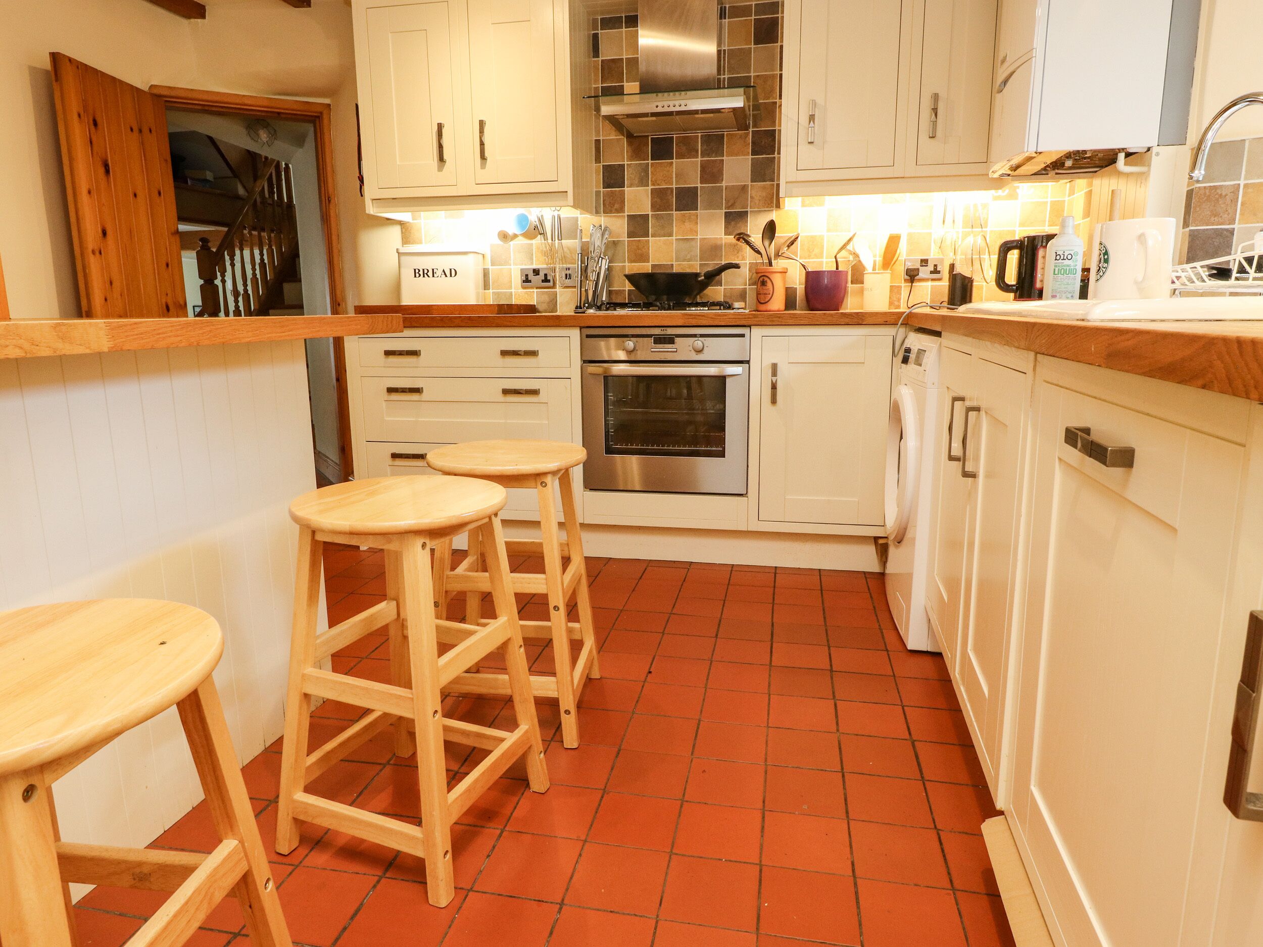 A kitchen with white cabinets, a stove, an oven, and a washing machine. There are three wooden stools and a countertop with various kitchen items.