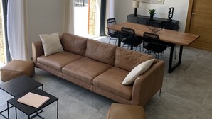 A modern living room with a brown leather sofa, a black coffee table, and a matching ottoman. A dining area with a wooden table and chairs is visible in the background.