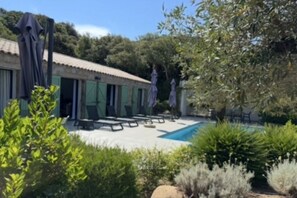 A pool area with lounge chairs and umbrellas, surrounded by greenery and a building with green shutters.