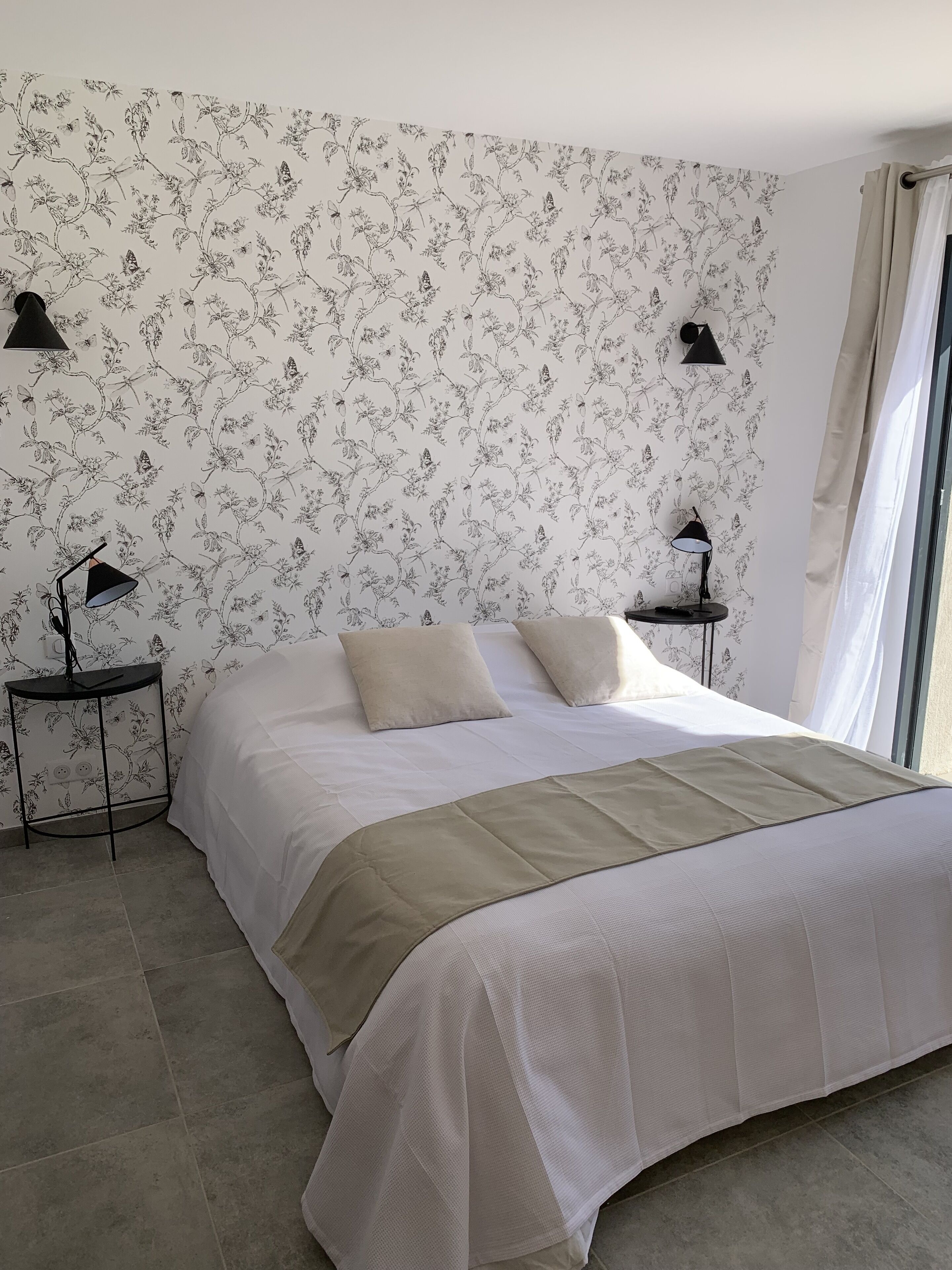 Ein Schlafzimmer mit einem großen Bett, zwei Nachttischen mit Lampen, blumigem Wandpapier und einem Fenster mit Vorhängen.