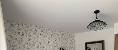 Una cama bien tendida con cabecera, una lámpara de noche, un aparador con cajones y un cuadro en la pared.