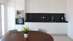 A modern kitchen with white cabinets, a black countertop, and stainless steel appliances. There is a dining table with chairs and a potted plant. - Nazaré Views I - Terrace, Pool & Sea View (Nazaré)